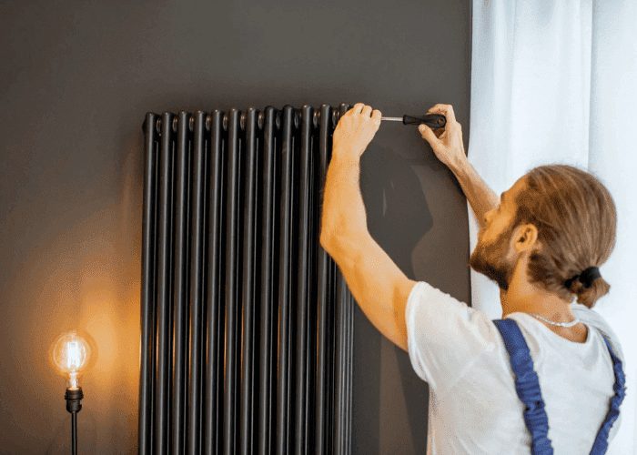 Technician in overalls using a screwdriver to adjust a tall black radiator on a dark wall.