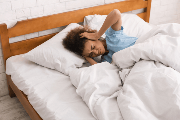 Woman lying in bed holding her head in discomfort after being disturbed by loud noise.