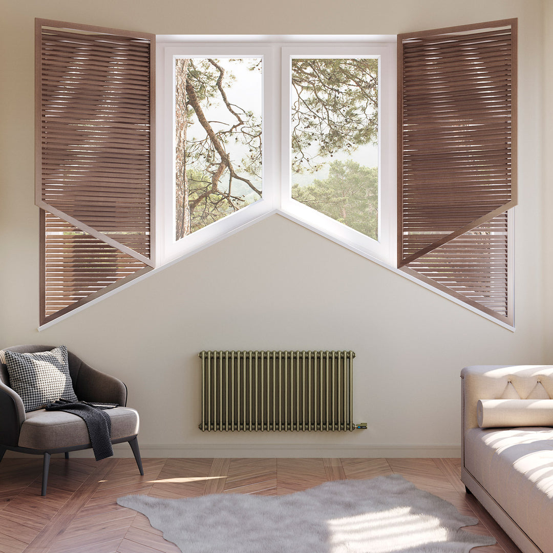 Anthracite designer radiator installed beneath a bay window in a bedroom with a bed, armchair and wooden shutters.