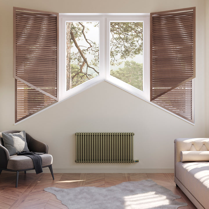 Anthracite designer radiator installed beneath a bay window in a bedroom with a bed, armchair and wooden shutters.